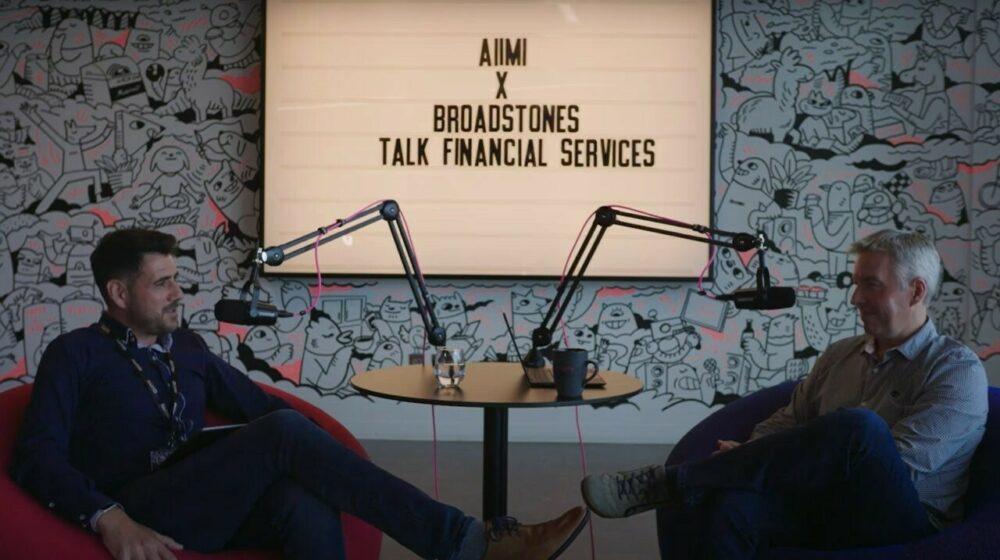 Two men sit in armchairs with microphones, discussing financial services. A sign behind them reads "AIIMI X Broadstones Talk Financial Services."