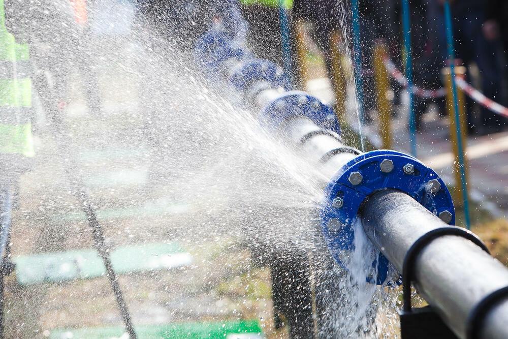 Water spraying from a large pipe with blue flanges, indicating a leak or burst under pressure in an outdoor setting.