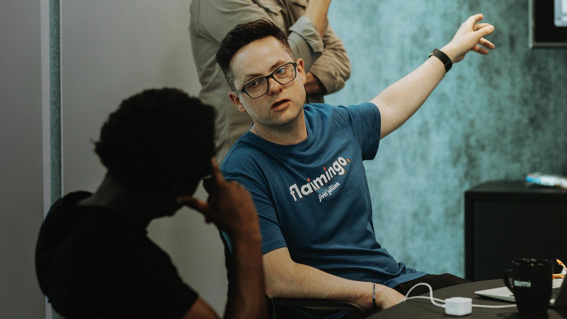 Man in glasses and blue shirt gestures while talking to another seated person in a casual meeting setting.