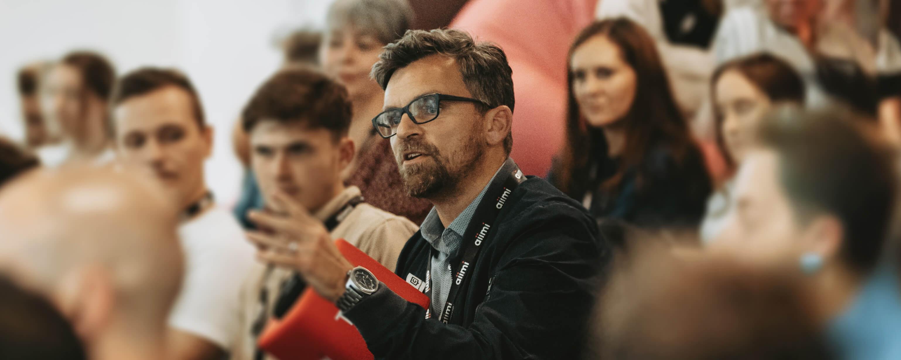 A man with glasses speaks in a group setting, holding a red folder. Several people are seated around him, attentively listening.