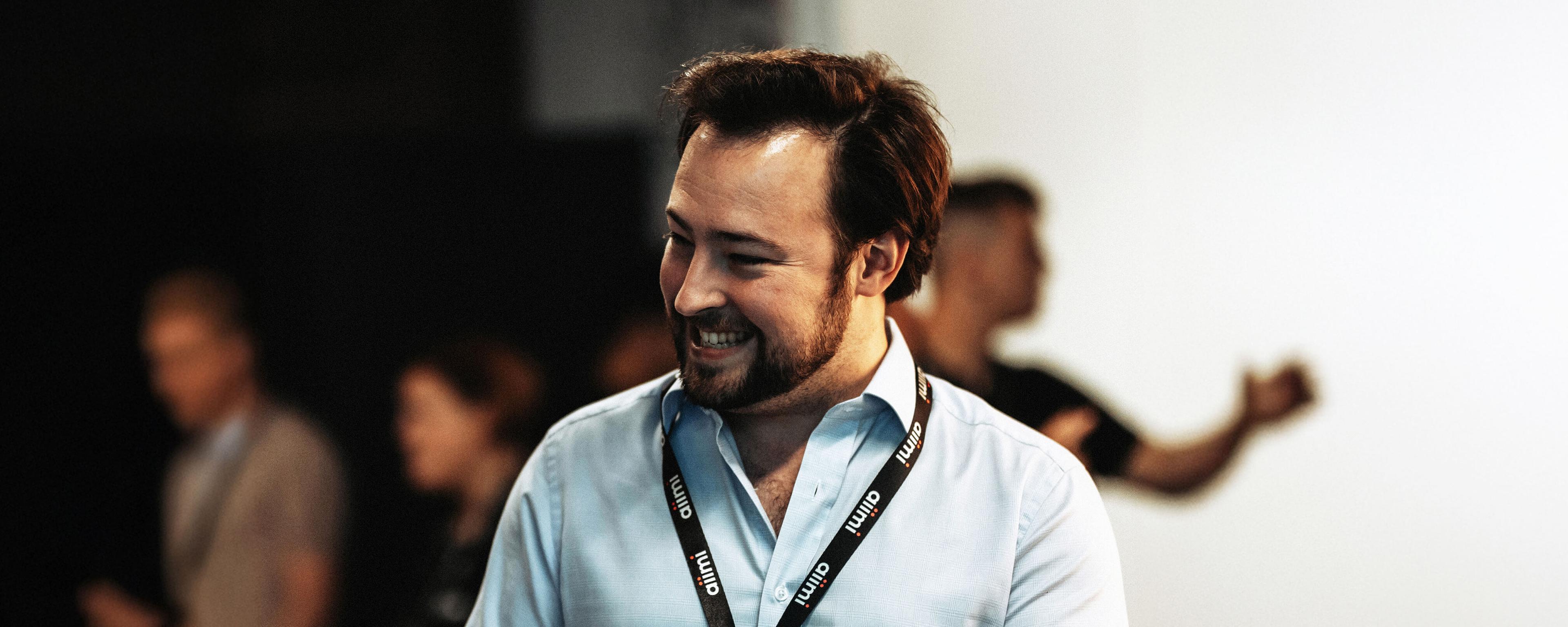 A man wearing a light blue shirt and a lanyard smiles while standing indoors. Blurred people are in the background.