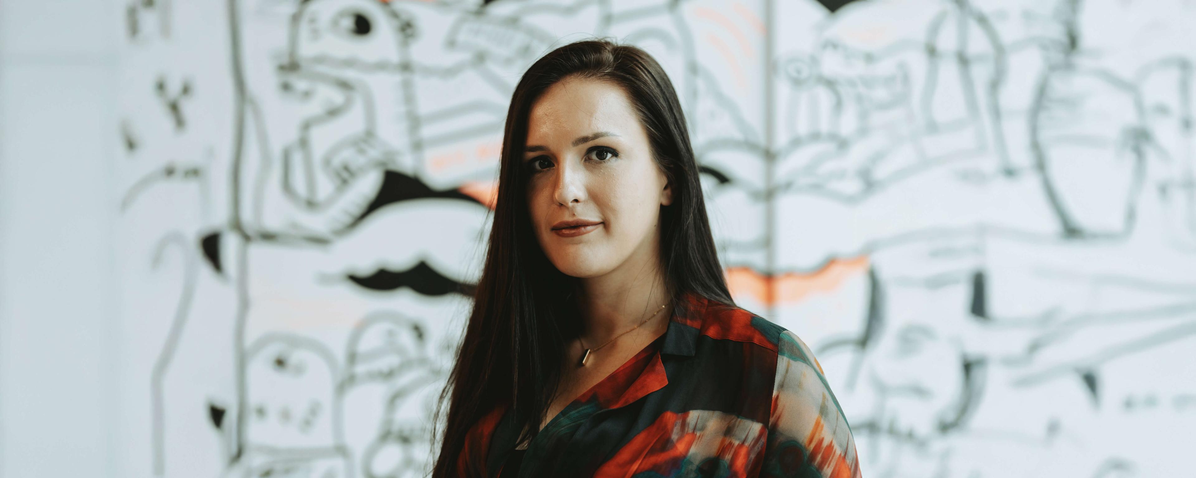Woman with long hair in a colorful blouse stands in front of a wall with abstract black and white art.