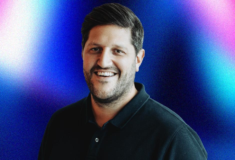 Smiling man with short dark hair and beard, wearing a black polo shirt against a vibrant blue and pink gradient background.