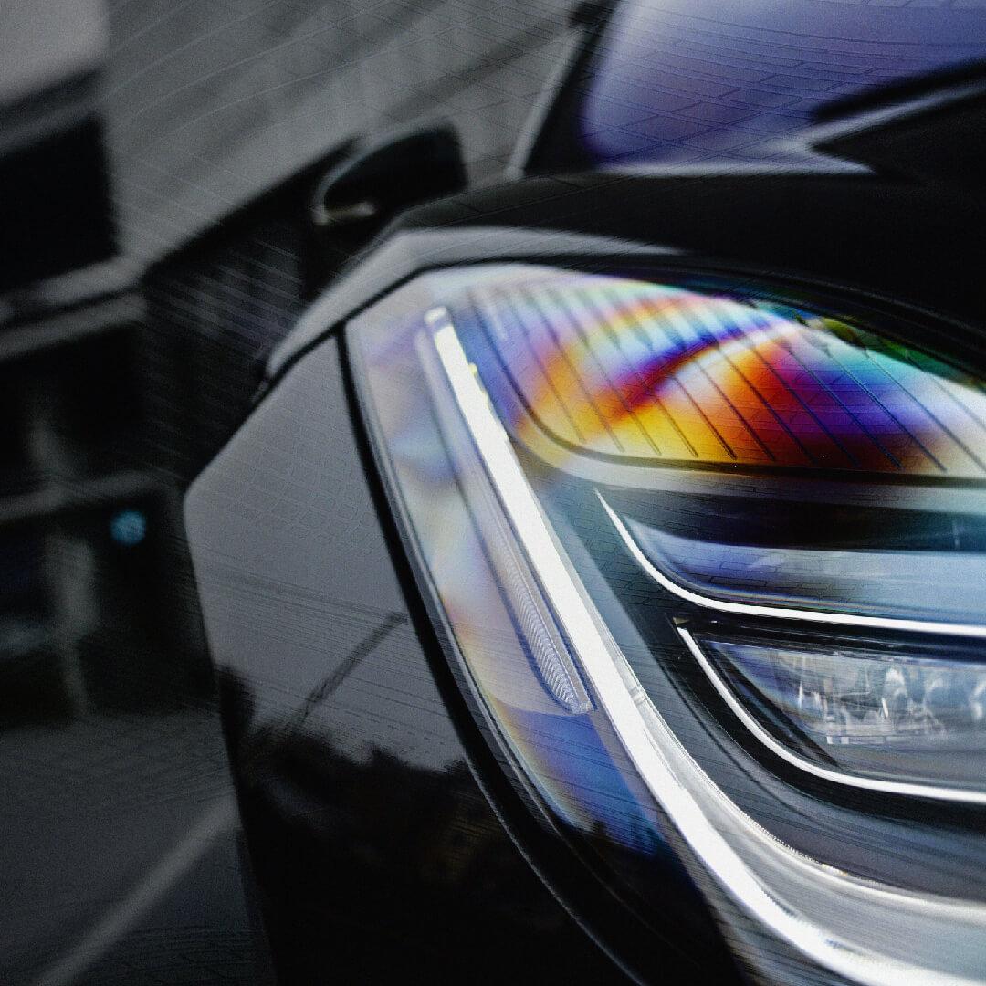 Close-up of a car's sleek headlight with a rainbow-like reflection on the glass surface, set against a blurred urban background.