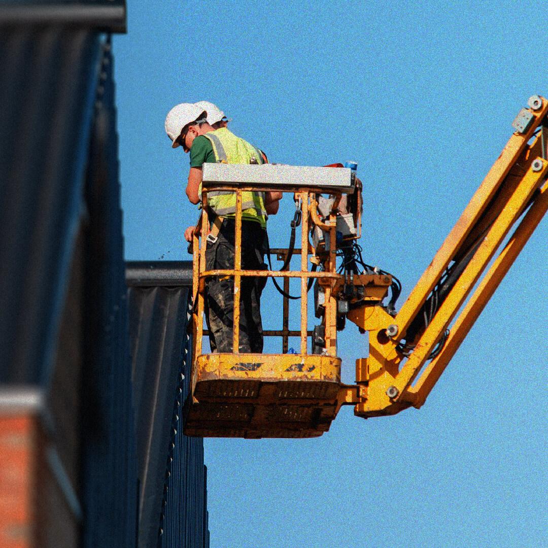 Two construction workers in safety gear on a yellow lift, working on a building facade against a clear blue sky.