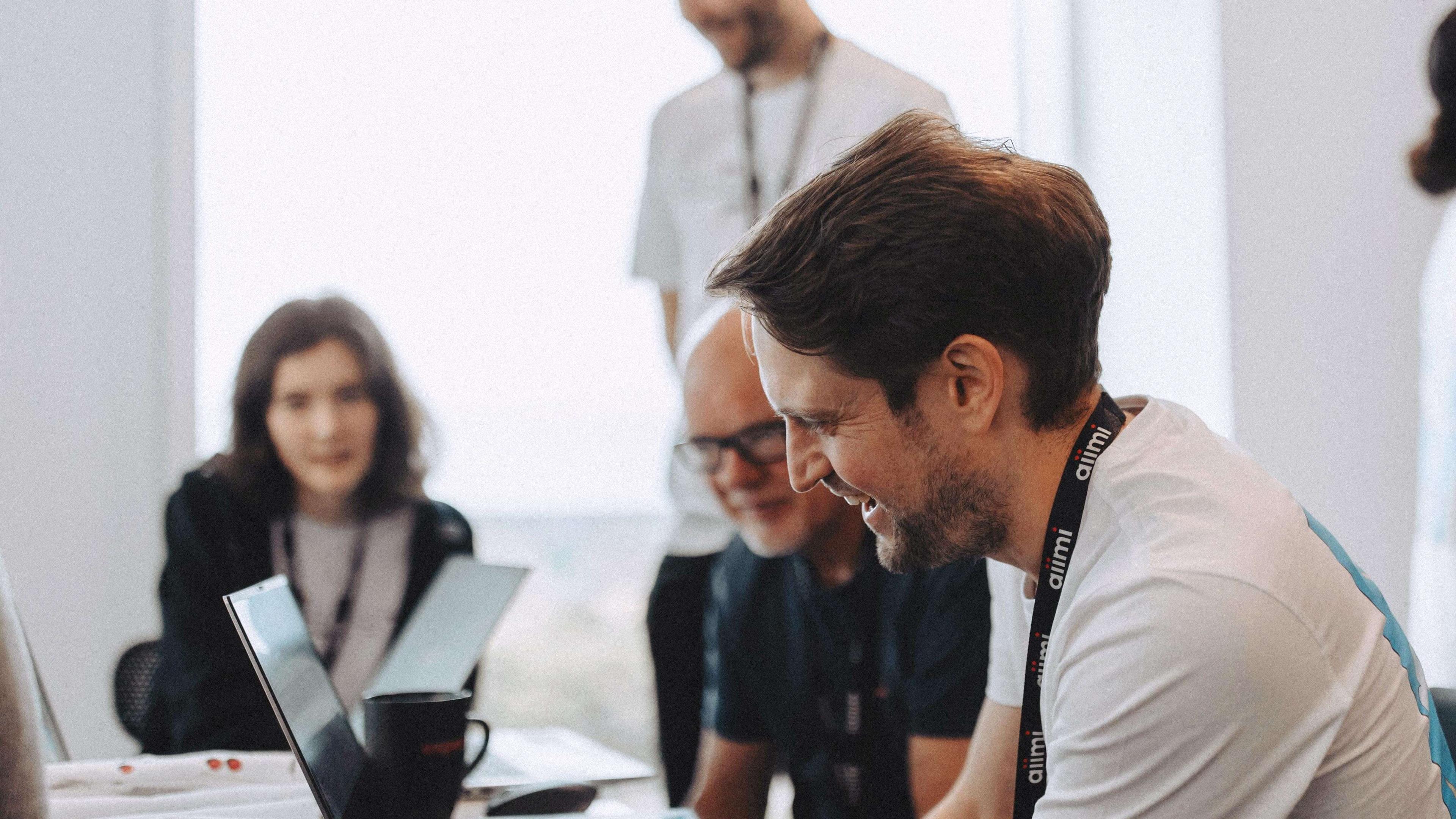 Smiling man with a lanyard works on a laptop, surrounded by colleagues in a bright office setting.